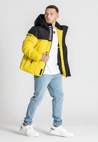 Gul og sort puffjakke med hætte, med lynlås foran og logo detaljer på kraven, båret over en hvid skjorte og blå jeans.
