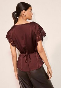 Top din satin în nuanța burgundy, cu mâneci din dantelă, având un talie legată și o siluetă fluidă. Textură netedă, detalii de design elegante la mâneci.