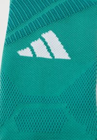 Meia teal feita de material de malha texturizada com um logótipo triangular branco. Apresenta um padrão de linhas diagonais e acentos pontilhados.