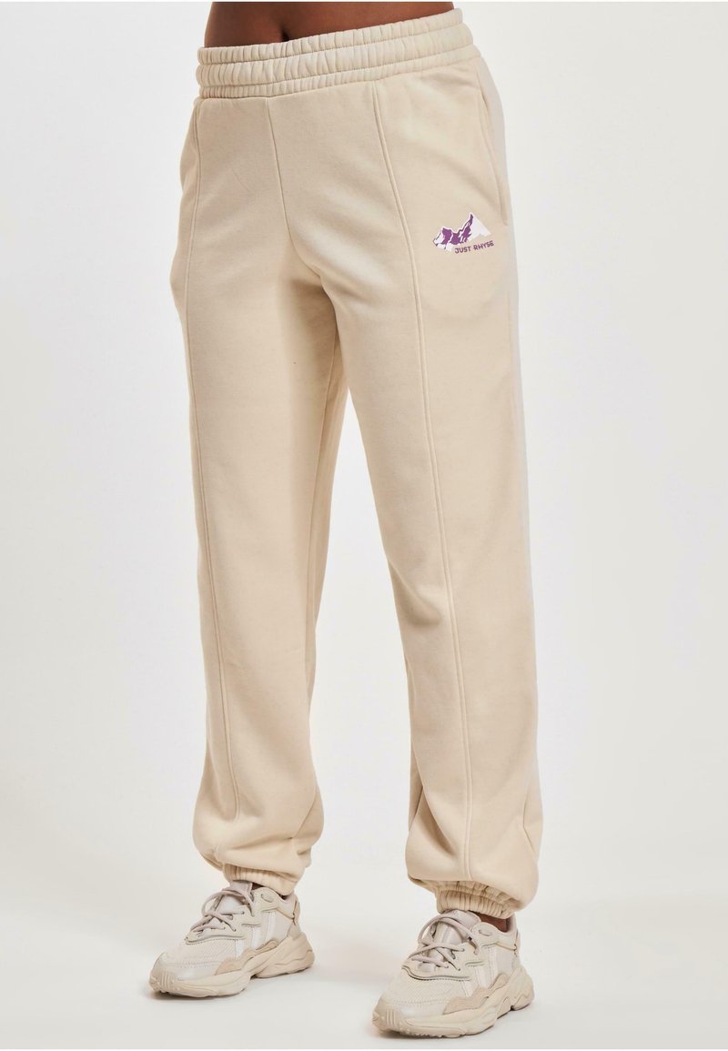 Beige Jogginghose aus weichem Stoff, mit einem elastischen Taillenbund, seitlichen Taschen und einem kleinen lila Logo auf dem Oberschenkel.