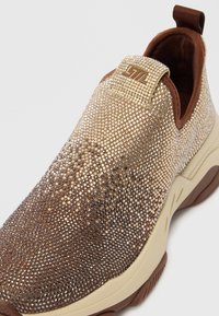 Slip-on stila sporta kurpes ar brūnu auduma oderi, krēmkrāsas zoli un augšdaļu, kas pārklāta ar zelta un sudraba rhinestones, ar zīmola cilpu vilkšanai.