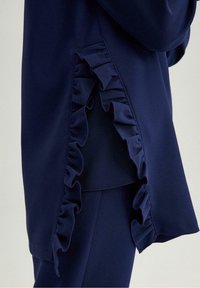 Marineblauwe top met een rimpeldetails aan de zijkant, gemaakt van een gladde stof. De rimpel voegt textuur toe langs de zoom.