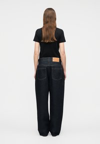 Mujer de cabello largo y castaño, vestida con una camiseta negra de manga corta y vaqueros de pierna ancha azul oscuro, de pie de espaldas sobre un fondo liso.