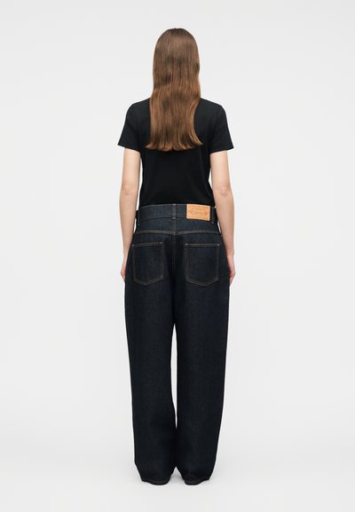 Femme aux longs cheveux bruns portant un t-shirt noir à manches courtes et un jean large bleu foncé, debout de dos sur un fond uni.