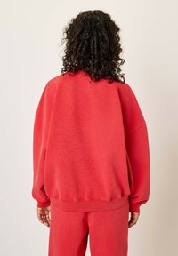 Sarkans pārmērīgs džemperis ar nolaižamām plecu šuvēm un ribotām piedurknēm. Mīksta tekstūra un brīva piegriezuma. Aizmugures skats rāda minimālistiskus dizaina elementus.