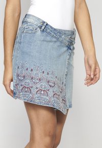 Denim krilo v svetlo modri barvi s cvetličnim vezenjem v rdeči in beli barvi ob robu. Ima zapiranje na gumb in stranske žepke.