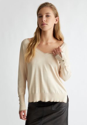 Maglione lavorato a maglia beige con scollatura a V, dettagli a onda e maniche lunghe con dettagli a bottoni ai polsini. Texture morbida.