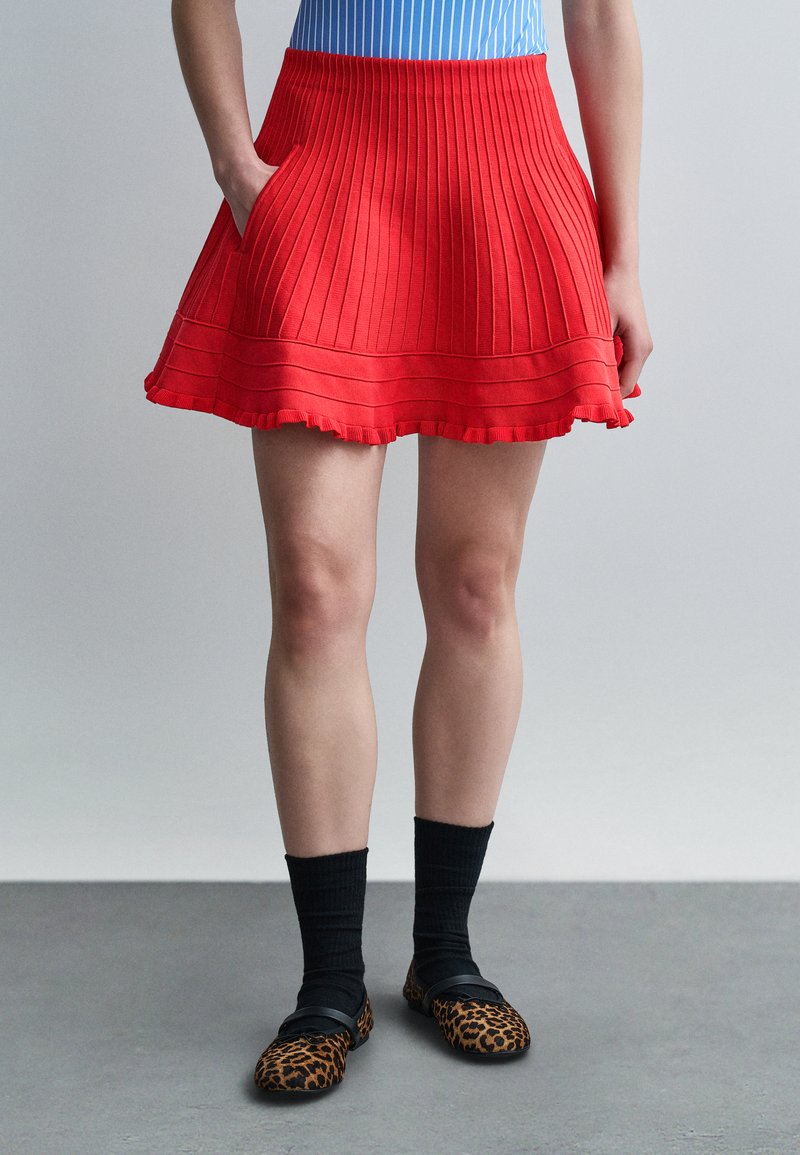 Femme portant une jupe plissée rouge avec des poches, des chaussettes noires et des chaussures plates à imprimé léopard, debout sur un sol gris devant un fond uni.