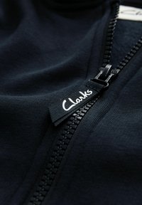 Sötétkék cipzáras dzseki sima szövettextúrával, látható cipzárral, műanyag fogakkal, és „Clarks” felirattal ellátott címkével.