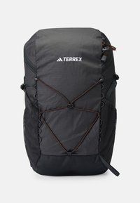 Mochila Terrex negra con una superficie texturizada, bolsillos laterales de malla y un acento de cordón elástico naranja. Presenta correas ajustables y un asa en la parte superior.