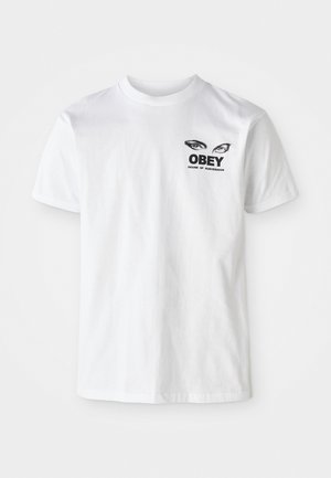 Balta kokvilnas T-kreklu ar melnu grafiku, kurā attēlotas acis un teksts "OBEY HOUSE OF SUBVERSION" uz krūtīm. Īsas piedurknes, apaļš kakla izgriezums.