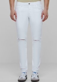 Blugi din denim alb, cu găuri distruse la genunchi, cu un design ajustat și stil standard cu cinci buzunare, asortați cu adidași albi.
