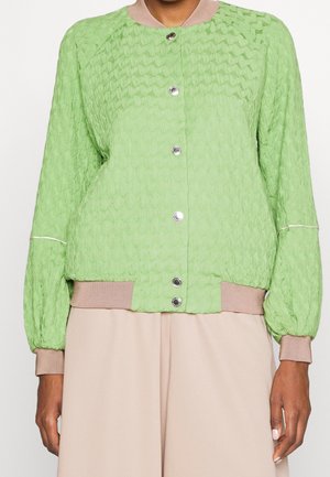 Chaqueta verde de manga larga con textura, con puños, dobladillo y cuello beige acanalados, con botones de presión plateados en la parte delantera, llevada sobre pantalones beige.