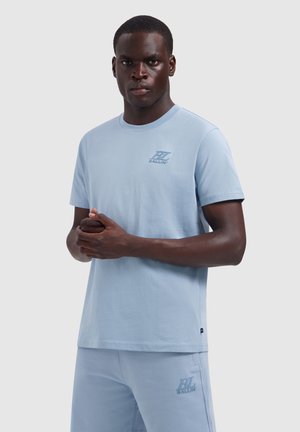 Jonge man met een donkere huid die een lichtblauw T-shirt en korte broek met een klein logo draagt, staat met gevouwen handen tegen een effen achtergrond.