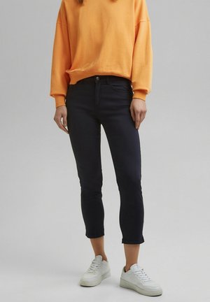 Personne portant un jean skinny noir raccourci, des baskets blanches et un haut décontracté à manches longues orange, debout devant un fond neutre.