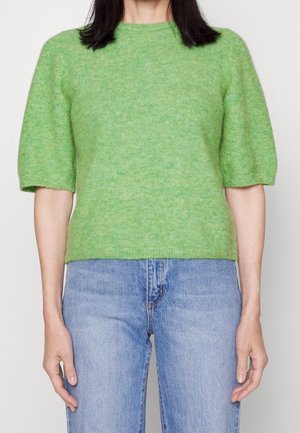 Person iført en lys grøn kortærmet strikket sweater og blå jeans, vist fra skuldrene til øvre lår, står mod en ensfarvet baggrund.