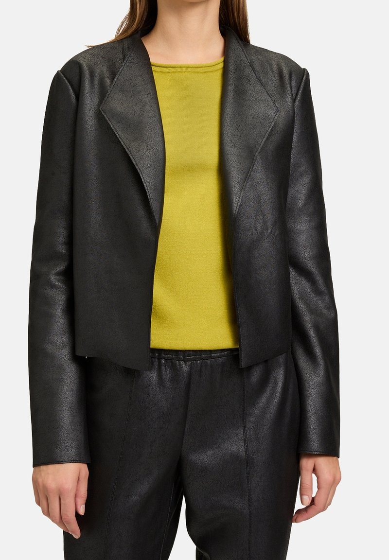 Femme portant une veste en cuir noire courte et un pantalon avec un haut jaune vif sur un fond uni.