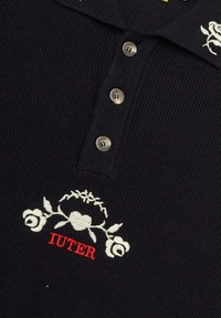 Polo nero a costine con tre bottoni metallici, design floreale ricamato in bianco e "IUTER" in rosso.