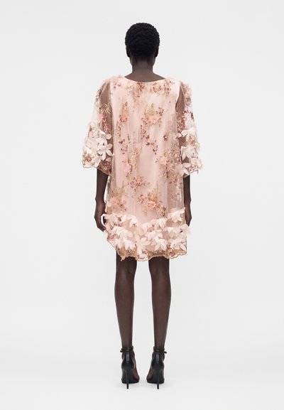 Γυναίκα φοράει ένα διαφανές ροζ φόρεμα με κεντήματα λουλουδιών και 3D εφαρμογές λουλουδιών, φαίνεται από πίσω, συνδυασμένο με μαύρα παπούτσια με ψηλό τακούνι.