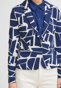 Blazer mariné et blanc à motifs en tissu lisse. Comprend des formes géométriques, une coupe ajustée, des revers crantés, et une fermeture par un seul bouton.