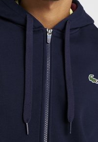 Tummansininen vetoketjullinen huppari, jossa on sileä kangas, punotut nyörit ja pieni vihreä krokotiililogo vasemmassa rintapielessä.