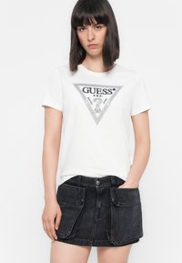Woman wearing white Guess T-shirt with black logo and black denim skirt with large front pockets, standing against a white background.