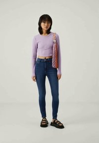 Top cropped de manga longa lilás claro, jeans skinny de cintura alta azul escuro e sandálias chunky pretas. Bolsa laranja e rosa com design em rede.