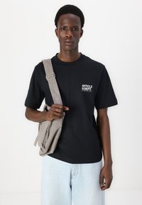 Black cotton t-shirt featuring a white logo on the chest, paired with light blue denim shorts and a grey crossbody bag.
