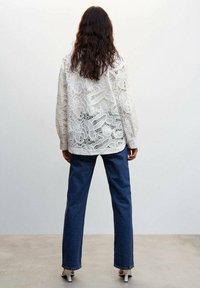 White lace blouse with intricate patterns, long sleeves, paired with denim trousers and high-heeled shoes, viewed from the back.
