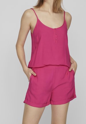 Kobieta w jaskrawym różowym topie bez rękawów z guzikami i pasujących spodenkach, z rękami w kieszeniach na jasnoszarym tle.