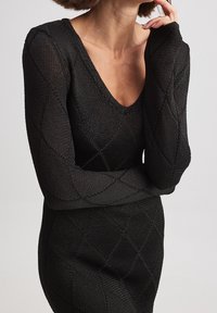 Femme portant une robe tricotée noire avec un décolleté en V profond et un motif en diamant, posant avec une main près de son visage et l'autre croisée.