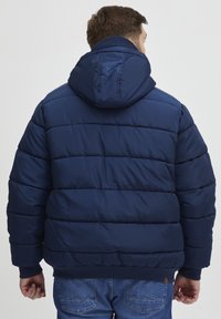 Tamsiai mėlynas pūkinis švarkas su kapišonu, turintis siūlomas quilted dalis ir riešo audinį. Medžiaga atrodo lygi ir izoliuota, puikiai tinkanti šaltam orui.