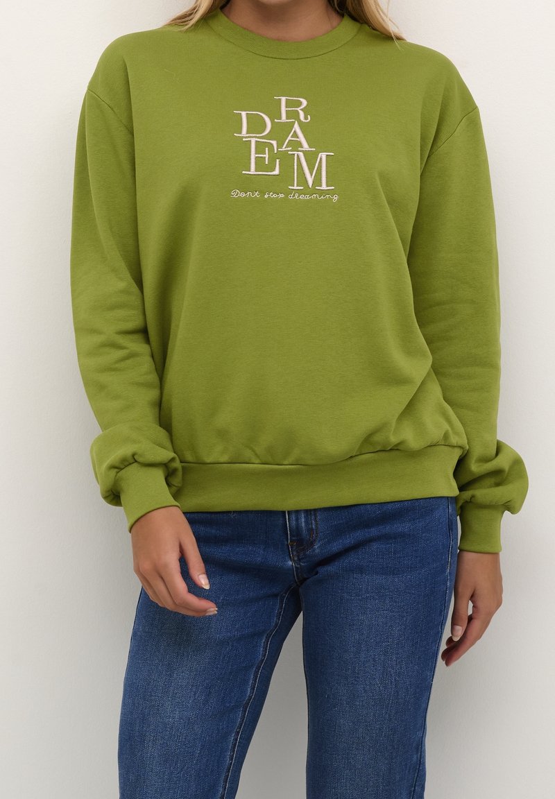 Groene sweatshirt gemaakt van een katoenmengsel, met "DREAM" geborduurd in lichtroze. Met boorden aan mouwen en zoom, gecombineerd met een blauwe spijkerbroek.