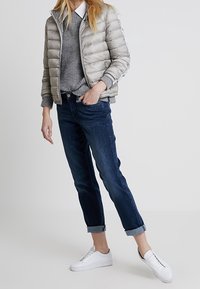 Daunenjacke in Hellgrau, gerippter grauer Pullover, dunkelblaue, hochgekrempelte Jeans und weiße Sneaker. Glatter, glänzender Stoff mit horizontalen Steppnähten.