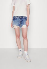 Henkilö, jolla on valkoiset lenkkarit, valkoiset sukat, revityt siniset farkkushortsit ja valkoinen t-paita, seisoo tasaisen valkoisen taustan edessä.