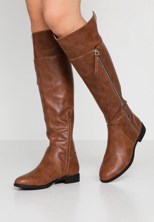 Bottes en cuir marron montant jusqu'aux genoux avec fermetures éclair latérales portées sur des jambes nues, debout sur un sol et un fond gris clair.