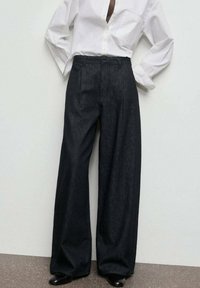 Pantalones de mezclilla de pernera ancha en azul oscuro con una cintura alta, textura suave y plisados en la parte delantera, combinados con una camisa blanca de botones.
