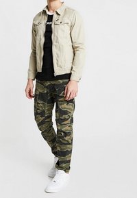 Mężczyzna nosi beżową kurtkę na czarnej koszulce, spodnie typu cargo w kamuflażu oraz białe trampki, stoi na tle gładkiego, białego tła.