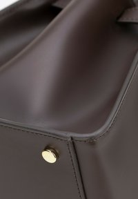 Brązowa skórzana torba o zorganizowanej konstrukcji; posiada złoty metalowy akcent oraz widoczne szwy. Gładka tekstura i czyste linie.