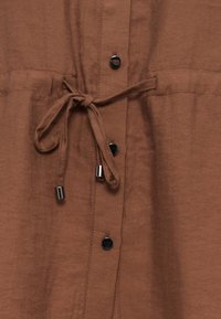 Καφέ ύφασμα με εμπρόσθια μεταλλικά κουμπιά και κορδόνι δέσης με μεταλλικά άκρα.