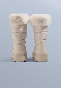 Beige waterdichte laarzen met een zachte witte bontafwerking aan de bovenkant, een gladde synthetische upper en een dikke, gestructureerde zool voor extra grip.