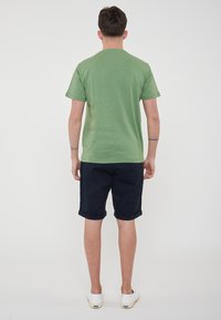 T-shirt verde a maniche corte realizzato in tessuto morbido, abbinato a pantaloni corti blu navy arricciati. Il modello indossa scarpe bianche senza lacci. Sfondo neutro.