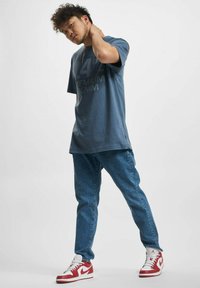 Blå grafisk t-skjorte med teksten "14% Premium Denim", kombinert med lyseblå jeans og røde/hvite joggesko. Herlig passform, avslappet stil.