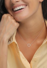 Srebrna ogrlica s obeskom v obliki deteljice, okrašenim z majhnimi prozornimi kamni, nošena prek svetlo rumene srajce z ovratnikom.