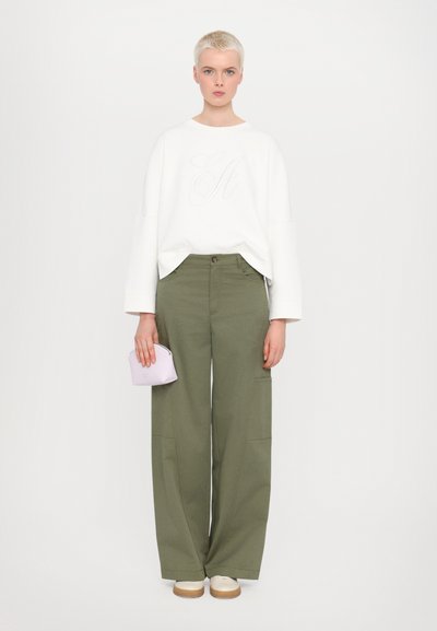 Sudadera blanca con gran logo bordado de 'G', pantalones wide-leg verde oliva con bolsillos laterales, combinados con zapatos beige y un bolso lila.
