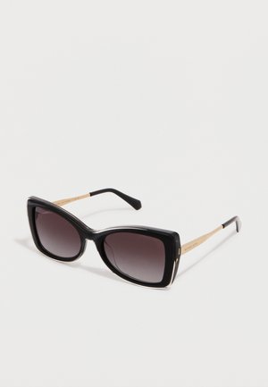 Lunettes de soleil œil de chat noires avec des verres dégradés foncés et des branches fines dorées posées sur une surface blanche.