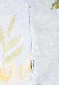 Un tissu bleu clair présente des motifs de feuilles jaunes et vertes, avec une texture visible et une petite étiquette portant la mention "Nilla Bong."