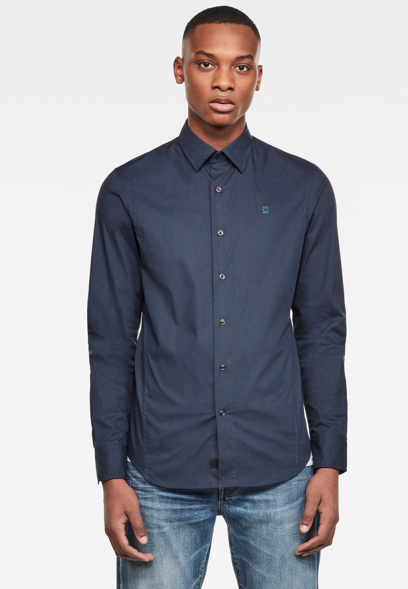 Camicia a maniche lunghe blu navy con chiusura a bottoni, colletto e logo blu sul lato sinistro del petto; abbinata a jeans in denim azzurri.