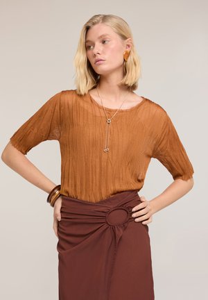 Vrouw met blond bobkapsel, draagt een gestructureerde gebrande oranje top, bruine rok met cirkelvormige ringdetail, gouden ketting en armbanden, poseert met handen in de heupen.