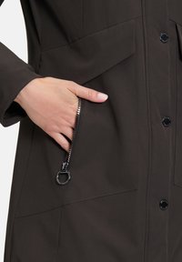 Veste marron foncé avec une poche zippée dotée de fermetures en métal noir et d'un bouton. Texture lisse avec des détails de panneaux subtils.
